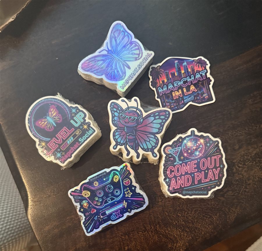 Stacks of holographic MapChat stickers on a wood table: the butterfly logo, MapChat in LA, Level Up, an XP controller, and Come Out and Play
